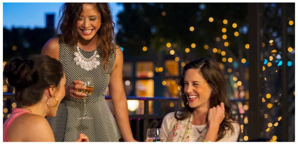Three women enjoying drinks and conversation at an outdoor evening gathering with festive lights in the background.