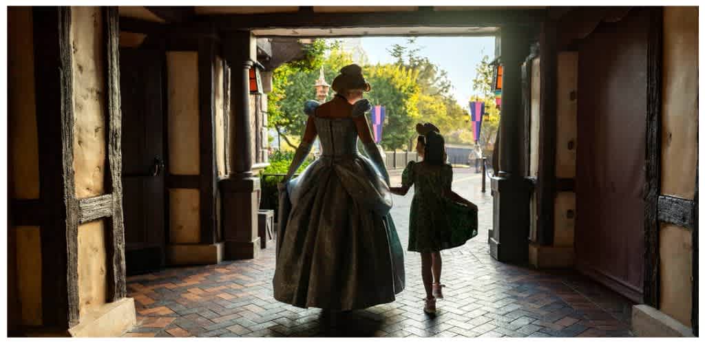 A woman and a girl holding hands walk out of a rustic building towards a sunny outdoor area with trees and colorful banners.