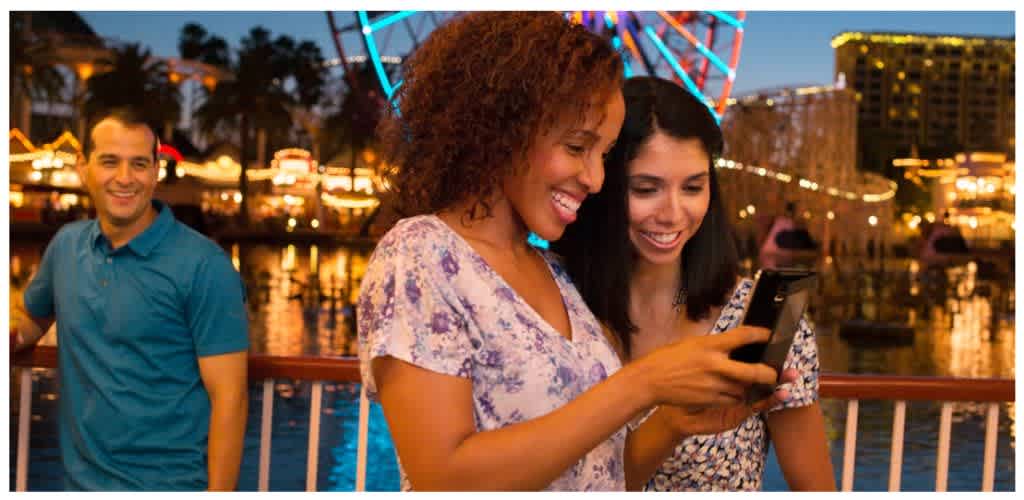 Two women smile and look at a smartphone together at an outdoor waterfront amusement park during evening with lights and a ferris wheel in the background.