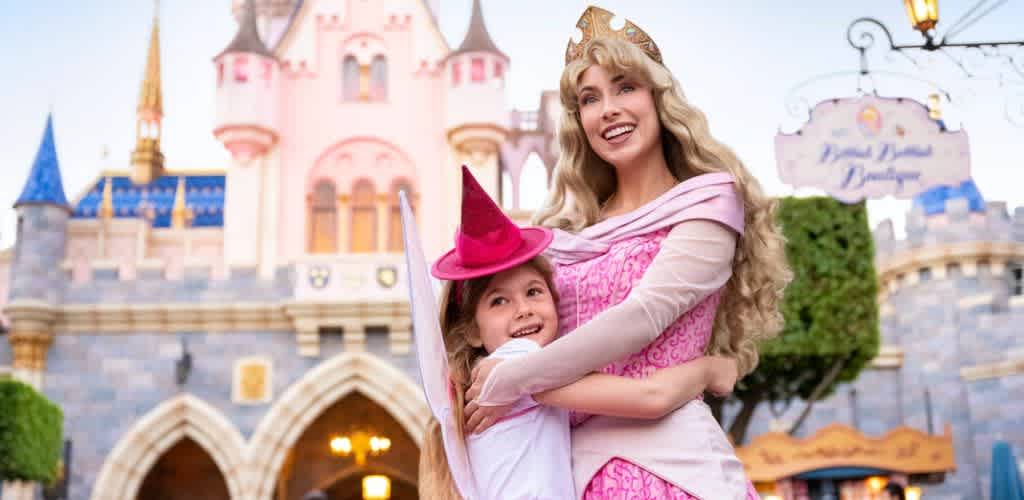A woman dressed as a princess hugs a girl in a pink hat in front of a castle at an amusement park.