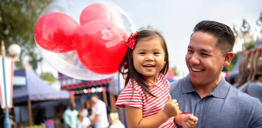 A smiling man holds a young girl with a red bow in her hair at an outdoor fair, with heart-shaped balloons and colorful tents in the background.