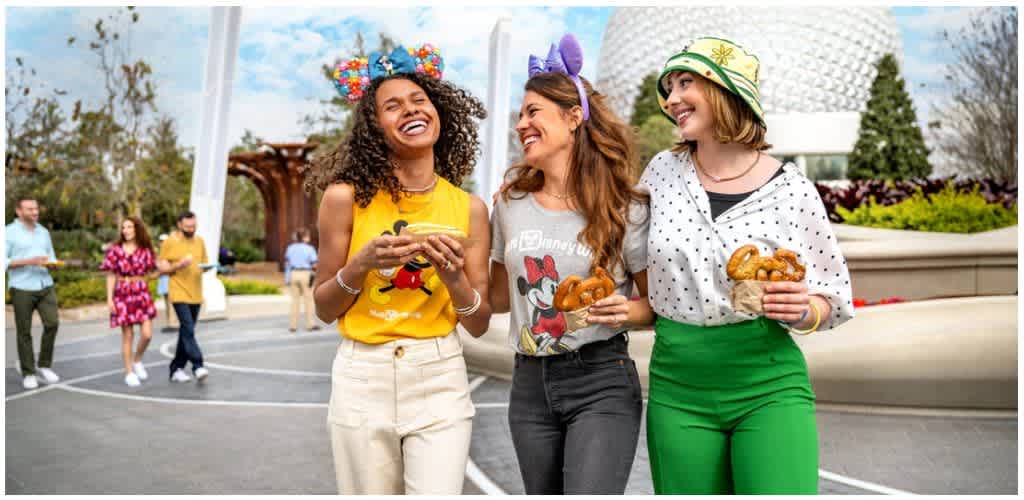 Three women smiling and holding pretzels outdoors during a festive event with people in the background and colorful decorations.