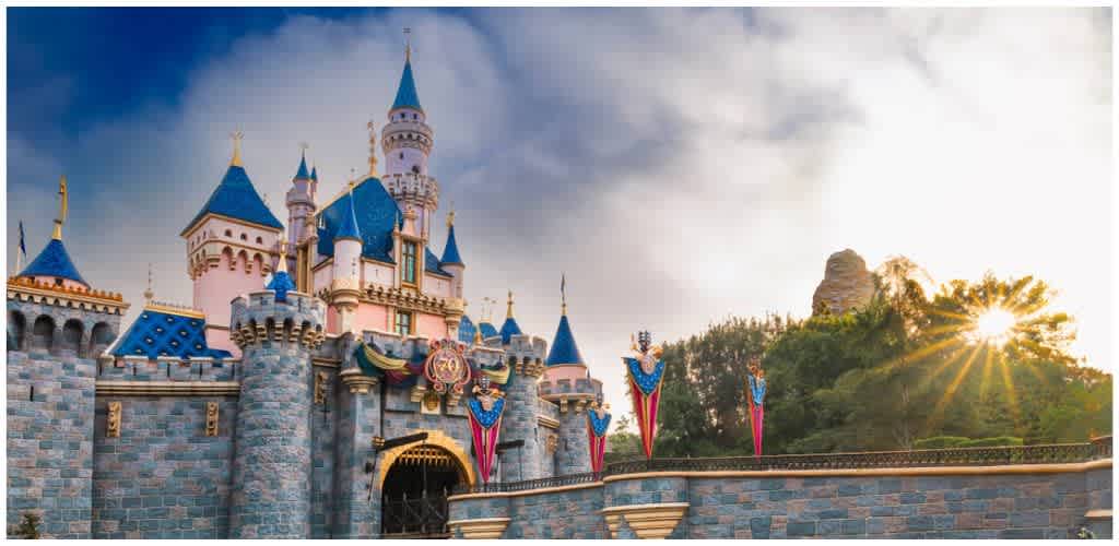 A colorful fairytale castle with blue rooftops, pink walls, and tall towers set against a bright sky with the sun shining through trees.