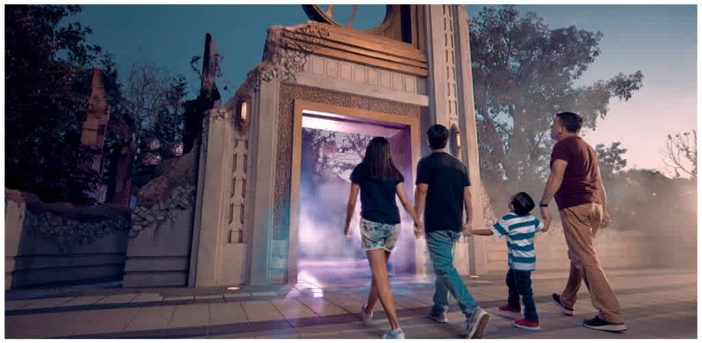 A family of four walking into a themed amusement park entrance with decorative architecture and atmospheric fog or smoke effects.