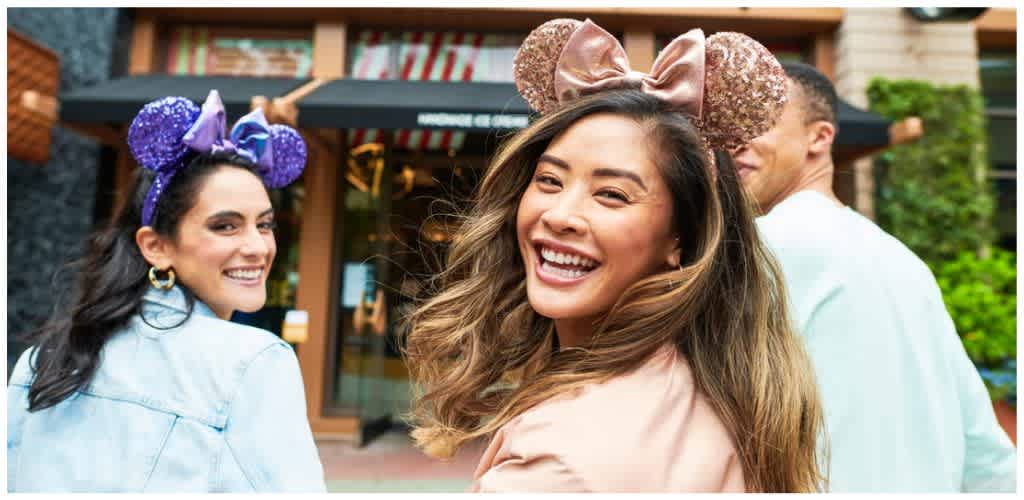 Group of smiling young adults wearing colorful Minnie Mouse ears outdoors with a background of storefronts and greenery