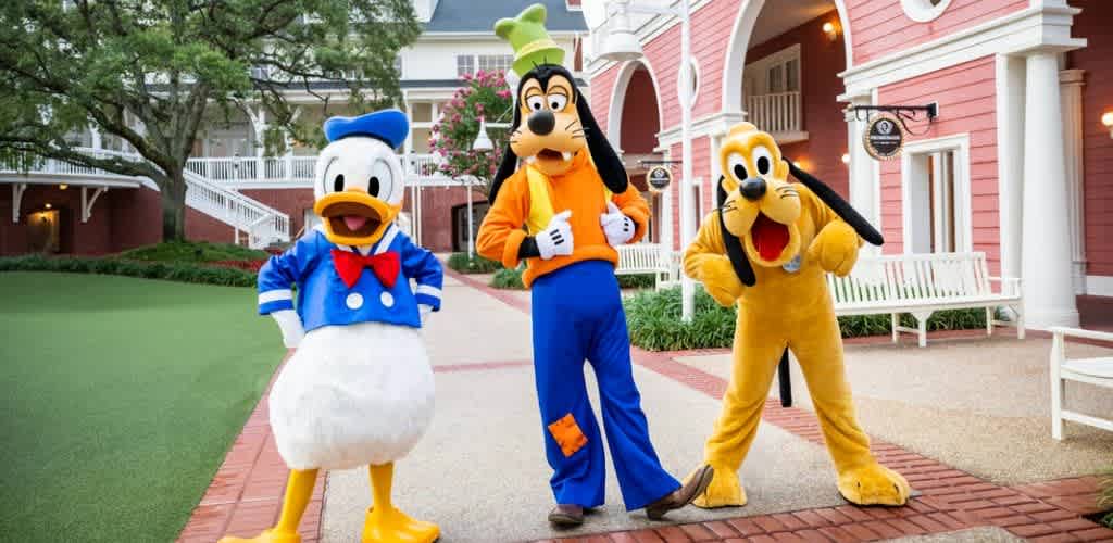 Costumed characters of Donald Duck, Pluto, and Goofy standing on a park pathway in front of a pink building with white accents and benches.