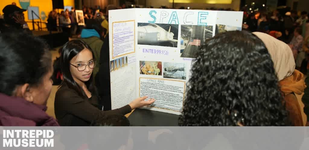 A woman presents information on a space-themed trifold board to a group of people at a museum exhibition.