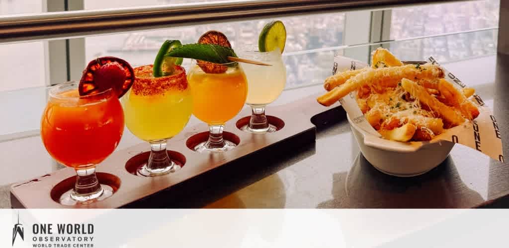 Four colorful cocktails on a wooden tray and a bowl of fries with cheese on a glossy surface near a window.