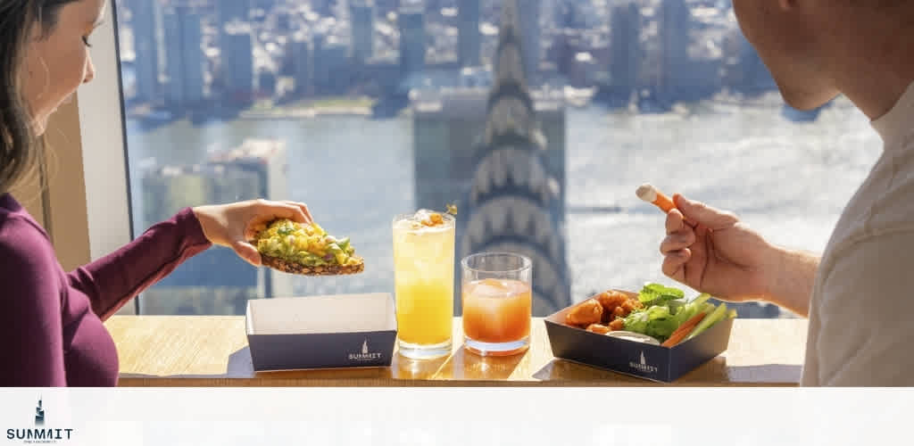 Two people share a meal with sandwiches and drinks at a table with a city skyline view through a large window. 