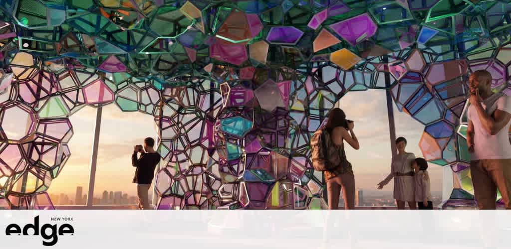 Colorful geometric glass sculpture with people inside and city view outside at sunset.