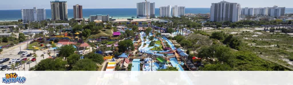 Aerial view of a colorful water park with rides, trees, and surrounding city buildings by the ocean