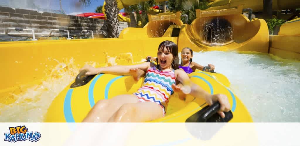 Two children enjoy riding a yellow water slide with water splashing around them at an amusement park