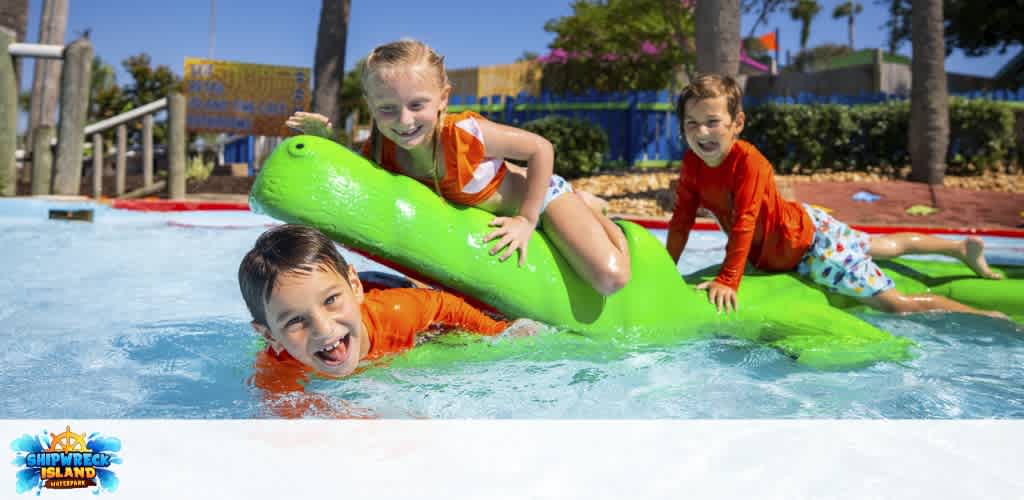 Three children playing on a green alligator-shaped water ride at Shipwreck Island Waterpark on a sunny day