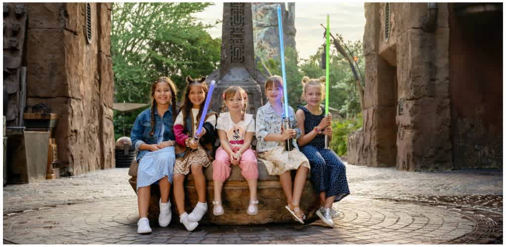 Five children sitting on a stone bench outdoors, holding colorful toy lightsabers, smiling at the camera amid natural and rocky surroundings.