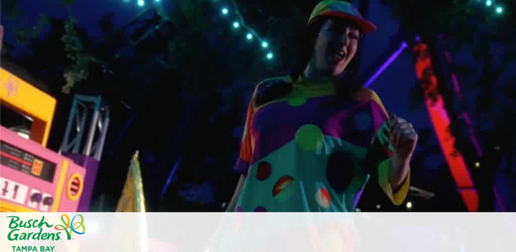 A performer in colorful costume with in bright lighting at an indoor amusement park or event space with colorful decorations and lights.
