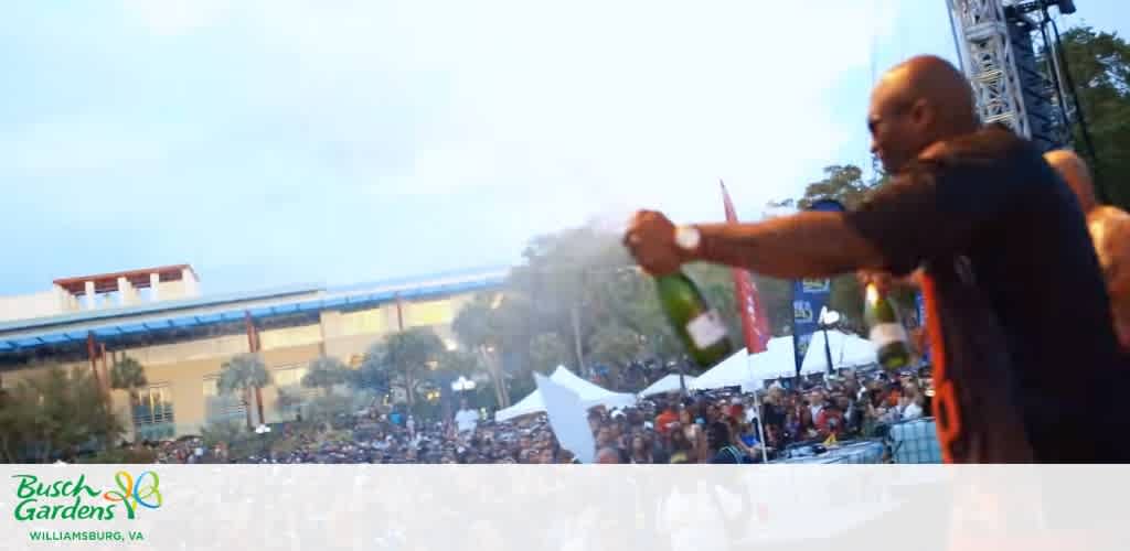 A person sprays champagne into the air at an outdoor event with many attendees, tents, and trees in the background at Busch Gardens Williamsburg Virginia