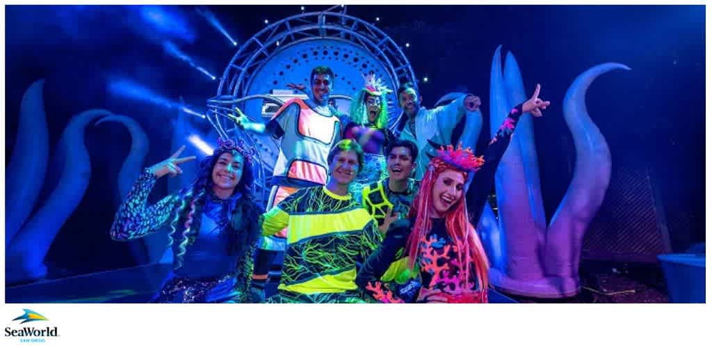 A group of performers in colorful, futuristic costumes pose energetically on an illuminated stage with tentacle-like structures.