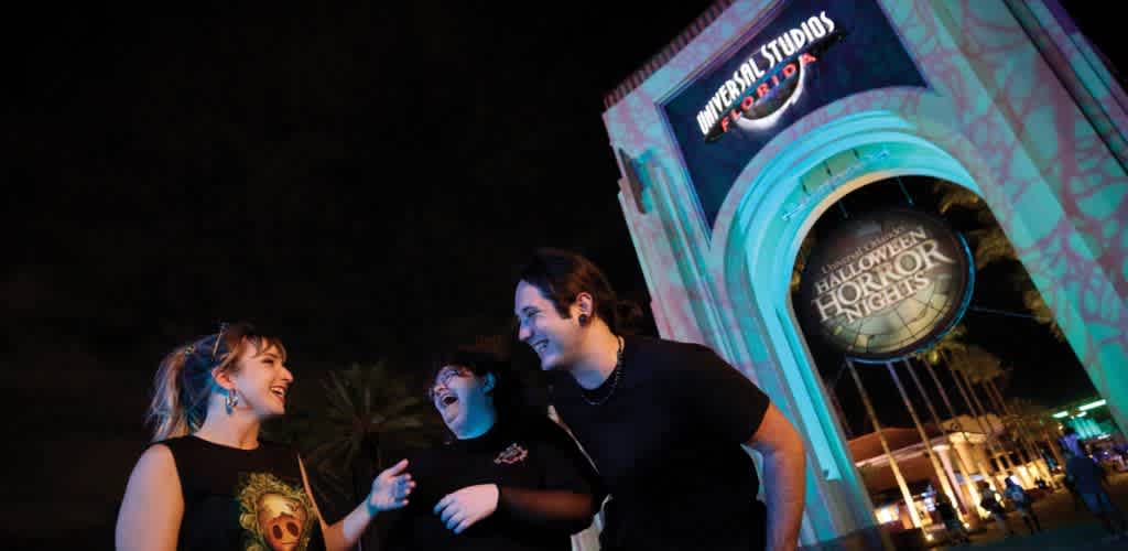 Three guests laughing together outside the glowing Halloween Horror Nights archway at the Universal Studios Florida entrance at night.
