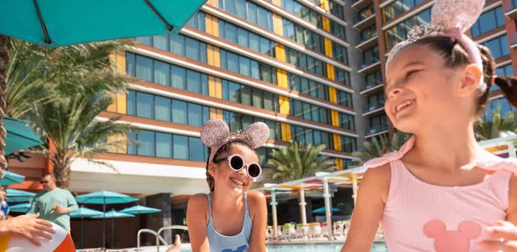 Two little girls wearing sparkly Mickey Mouse ears smiling and laughing poolside at a Disneyland Resort hotel on a sunny summer day.
