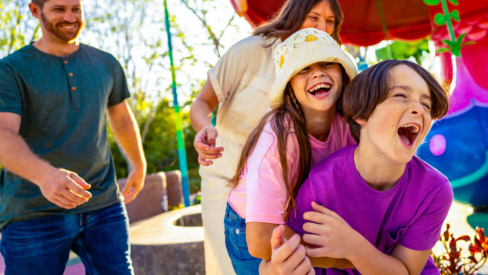 A family of four laughing and playing together in a brightly colored themed play area at the park.