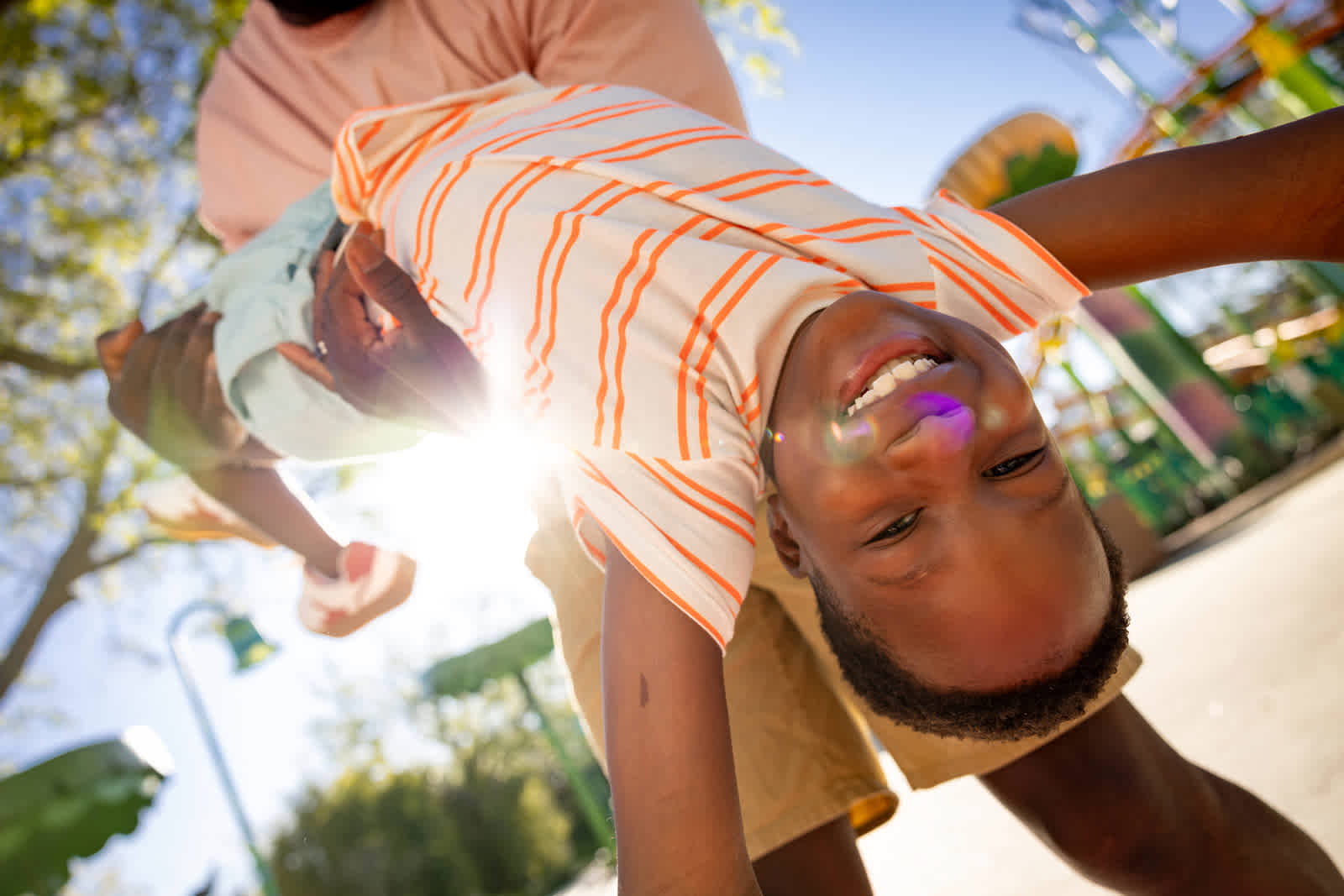A smiling child held up in the air by an adult at a sunny outdoor theme park surrounded by trees and colorful rides.