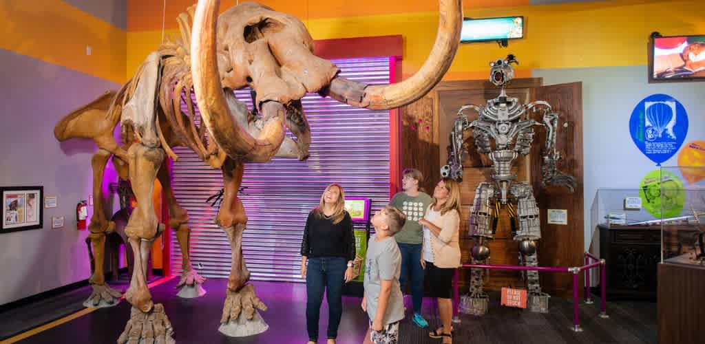 Group of people observing a large mammoth skeleton and a robot exhibit in a colorful museum setting