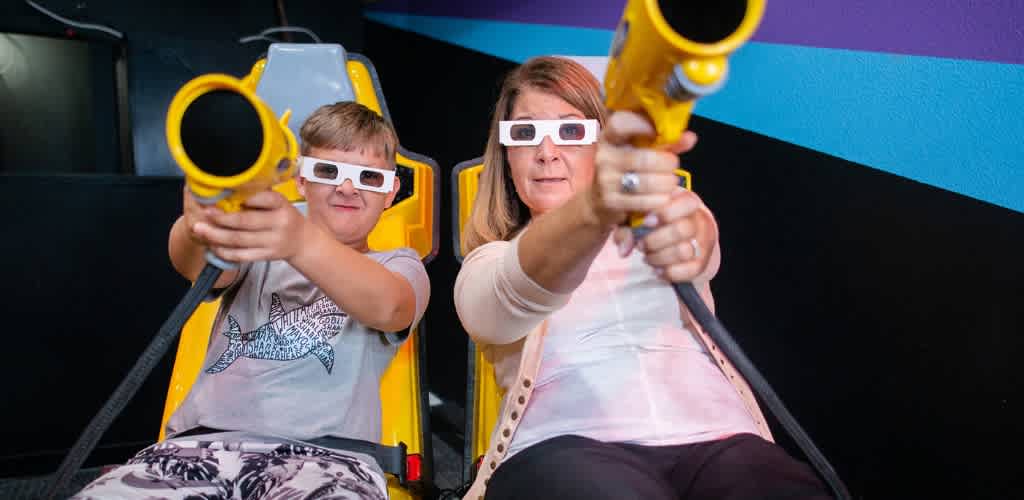 A boy and woman wearing 3D glasses and holding laser guns sit in yellow amusement ride seats ready for fun.