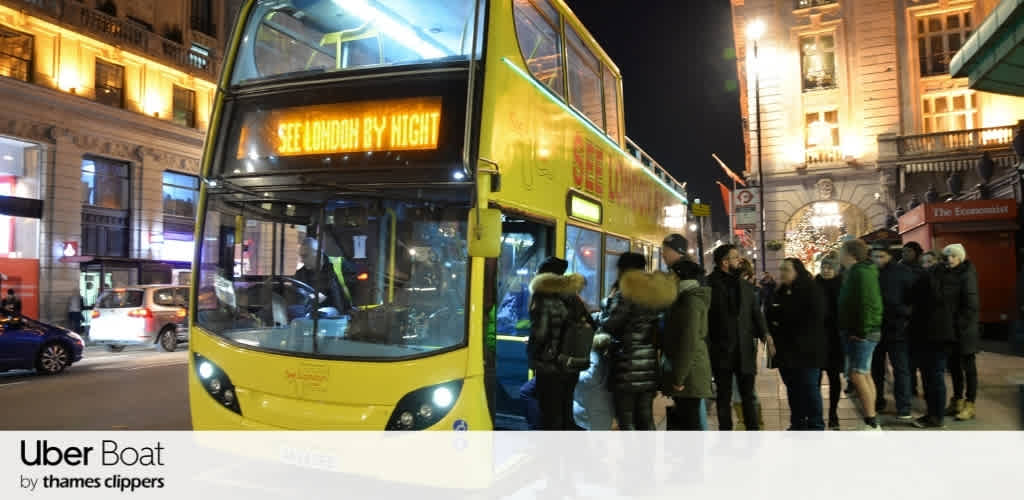 A double-decker yellow bus with passengers boarding at night in a busy city scene with illuminated buildings and a decorative archway in the background.