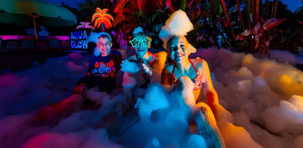 Three children play in vibrant foam at night, wearing colorful sunglasses and swimsuits