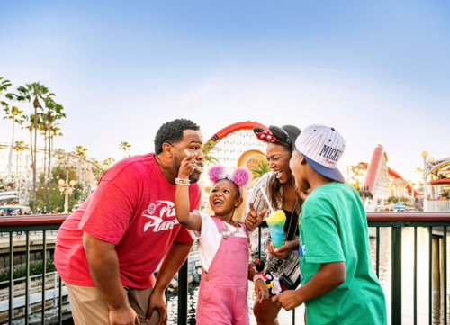 A family in red Disney shirts and Mickey ears laughing together on the Pixar Pier boardwalk at Disney California Adventure Park.