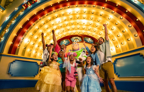 Children in princess dresses and casual outfits cheering with raised arms in front of a glowing marquee at Disneyland Park.
