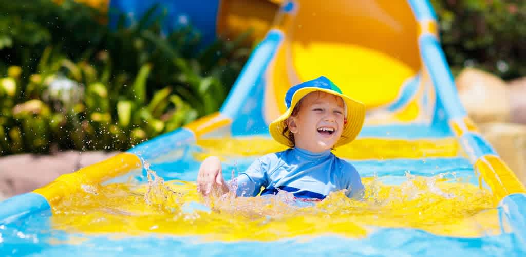 A happy child wearing a yellow and blue hat sliding down a water slide.