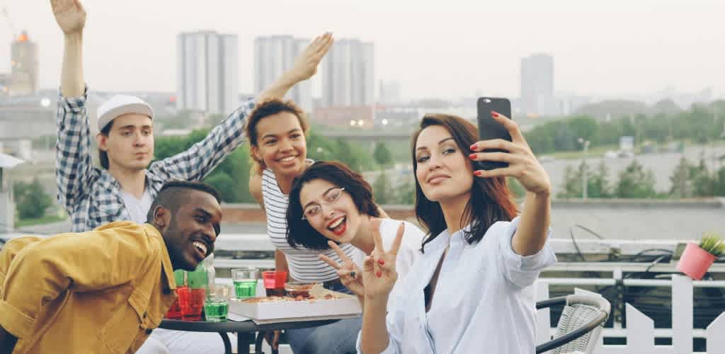 Group of five diverse friends taking a selfie on a rooftop terrace with city buildings in the background