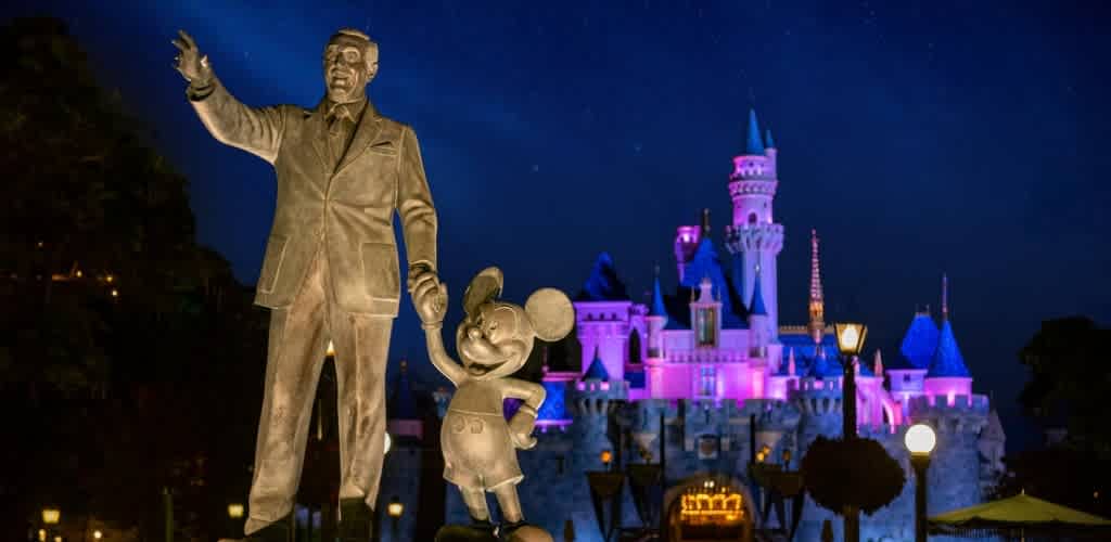 A nighttime view of a large statue of Walt Disney and Mickey Mouse in front of a colorful illuminated castle at Disneyland.