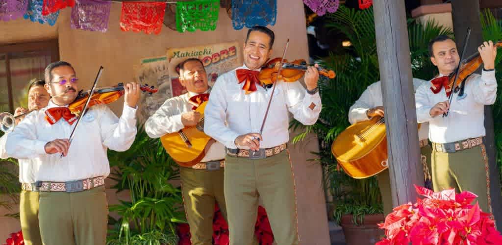 Group of musicians playing violins and guitars outdoors with colorful decorations and greenery in the background.