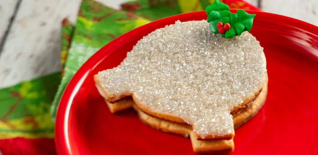 A sugar cookie shaped like a Christmas ornament with white sugar icing and green holly decoration on a red plate