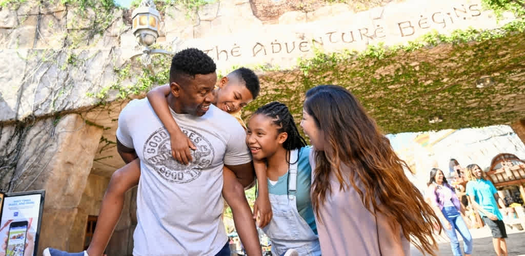 A family of four smiling and laughing together outdoors at an amusement park entrance. The background shows trees, rock formations, and other visitors enjoying the park.