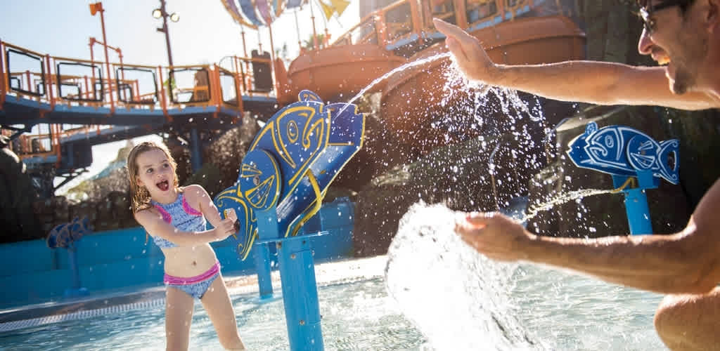 A young girl and an adult playing in a water splash area with colorful fish-themed water features and a water ride in the background