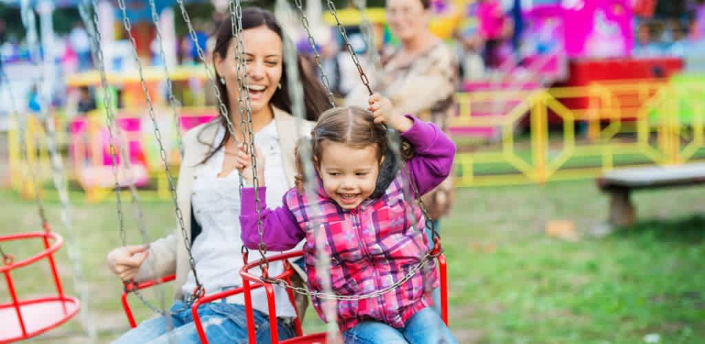 A smiling woman pushes a young girl on a colorful swing set outdoors on a sunny day at a playground.