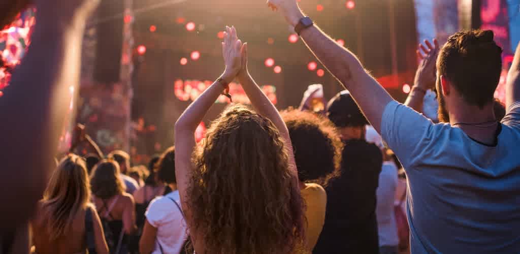 Crowd of people enjoying a concert outdoors with hands raised and stage lighting in the background