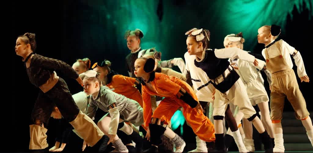 Children in colorful costumes perform on stage in a dynamic scene from a musical or play with a dark backdrop and dramatic lighting.