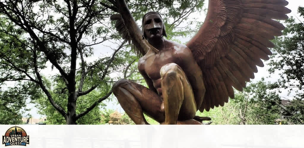 A bronze sculpture of a mythological figure with large wings and a mask, crouching outdoors among trees in a park setting
