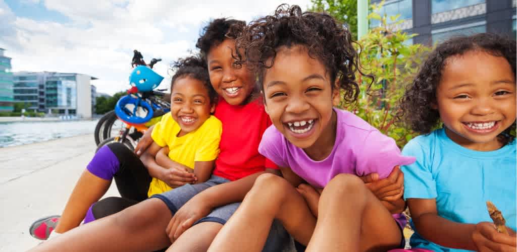 Group of children smiling and hugging outdoors near a bicycle and modern buildings on a bright day