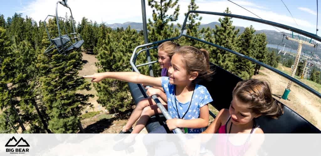 Three children enjoy a chairlift ride above a forested area. The scenery includes lush green trees under a clear blue sky, with mountainous terrain in the distance. The child on the left points outward, showing excitement at the view. The Big Bear logo is visible at the bottom-left corner, suggesting the location.