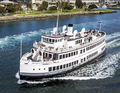 A vintage-style white ferryboat sailing with passengers, set against a backdrop of a waterfront and greenery.