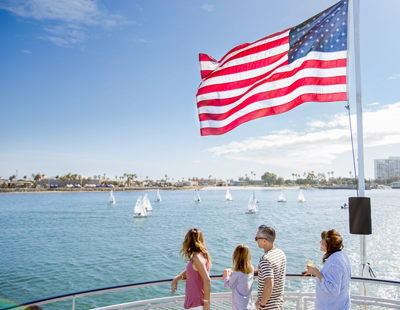 People on a dock watching sailboats near the American flag on a sunny day.