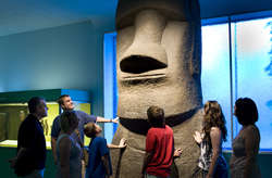 Visitors engage with a large Easter Island moai statue exhibit. A guide points to the towering figure while a diverse group, including children and adults, look on with interest. The exhibit room is softly lit, highlighting the statue's features and creating a contemplative atmosphere.