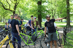 A group of cyclists wearing helmets pauses for instruction. A guide, labeled 'Unlimited Biking Guide', points out the route, while participants on bikes and one person standing look on, amid a backdrop of green trees in a park setting.