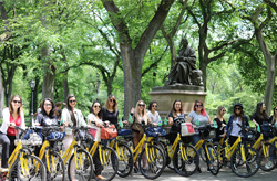 A diverse group of people is smiling and posing for a photo during an outdoor event. They're standing and kneeling on a bridge with lush trees in the background. Some individuals wave excitedly at the camera. Two yellow bicycles are prominently displayed, hinting at a cycling activity. Everyone appears to be in high spirits.