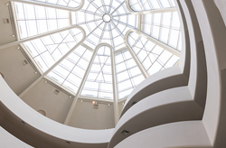 Image displays an architectural interior with white walls and a large domed skylight. The ceiling features geometric glass panes that allow natural light to flood the space. Below, broad, spiraling ramps create a sense of movement, guiding the eye towards the oculus at the top. No individuals are present in this serene, modern structure.